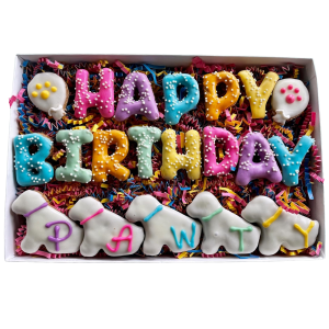 Image of a charming gift basket containing various handcrafted dog treats shaped like letters spelling 'HAPPY BIRTHDAY.' The basket is arranged with natural-looking, shredded paper filler and decorated with a bright, cheerful ribbon. The treats have a gol